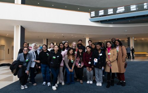 ALT text: A group of 20 students gather together, smiling, for a photo. Many are wearing coats and all have personal identification badges pinned to their clothing. 